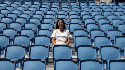 Smiling woman sitting alone in a mostly empty blue stadium seating area on a sunny day.
