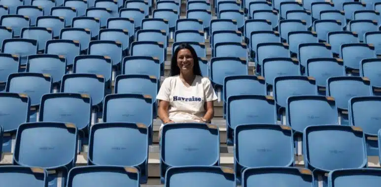 Screenshot | AllezPaillade.com Smiling woman sitting alone in a mostly empty blue stadium seating area on a sunny day.