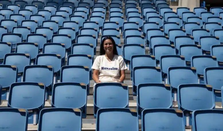 Smiling woman sitting alone in a mostly empty blue stadium seating area on a sunny day.