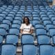 Smiling woman sitting alone in a mostly empty blue stadium seating area on a sunny day.