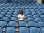 Smiling woman sitting alone in a mostly empty blue stadium seating area on a sunny day.