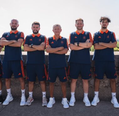 Seven men in matching navy and orange athletic uniforms pose with arms crossed along a stone wall outdoors.