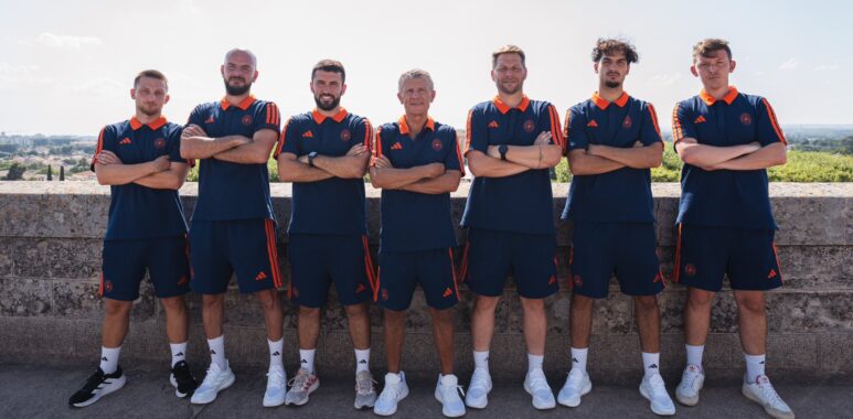 Seven men in matching navy and orange athletic uniforms pose with arms crossed along a stone wall outdoors.