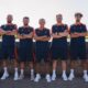 IMG_5452 | AllezPaillade.com Seven men in matching navy and orange athletic uniforms pose with arms crossed along a stone wall outdoors.