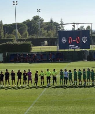 Lineups of two soccer teams shaking hands before a match on a sunny field, scoreboard showing 0-0 in the background