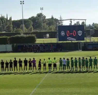 Lineups of two soccer teams shaking hands before a match on a sunny field, scoreboard showing 0-0 in the background