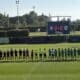 Lineups of two soccer teams shaking hands before a match on a sunny field, scoreboard showing 0-0 in the background