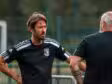 Two men in black athletic shirts stand and talk on a sports field, one facing the camera with hands on hips.