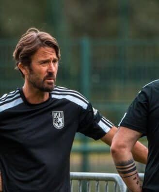 Two men in black athletic shirts stand and talk on a sports field, one facing the camera with hands on hips.