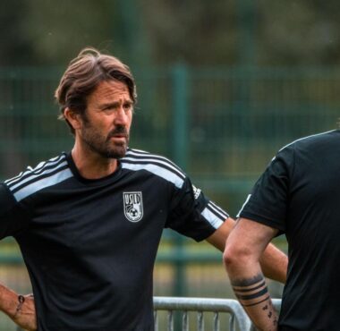 Two men in black athletic shirts stand and talk on a sports field, one facing the camera with hands on hips.