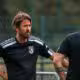Two men in black athletic shirts stand and talk on a sports field, one facing the camera with hands on hips.