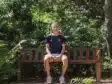 Person sits on a wooden park bench surrounded by lush green foliage, wearing a navy athletic shirt and light shorts.