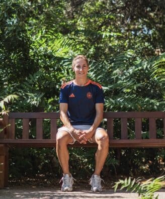 Person sits on a wooden park bench surrounded by lush green foliage, wearing a navy athletic shirt and light shorts.