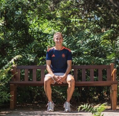 Person sits on a wooden park bench surrounded by lush green foliage, wearing a navy athletic shirt and light shorts.
