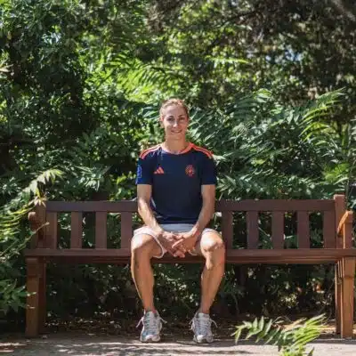 Person sits on a wooden park bench surrounded by lush green foliage, wearing a navy athletic shirt and light shorts.