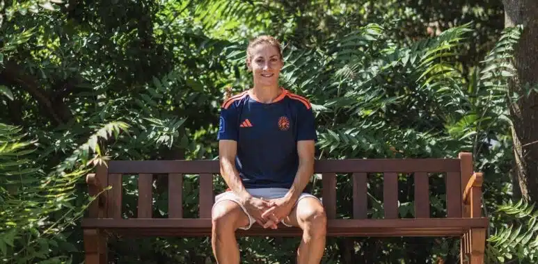 Person sits on a wooden park bench surrounded by lush green foliage, wearing a navy athletic shirt and light shorts.