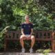 Person sits on a wooden park bench surrounded by lush green foliage, wearing a navy athletic shirt and light shorts.