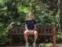 Person sits on a wooden park bench surrounded by lush green foliage, wearing a navy athletic shirt and light shorts.