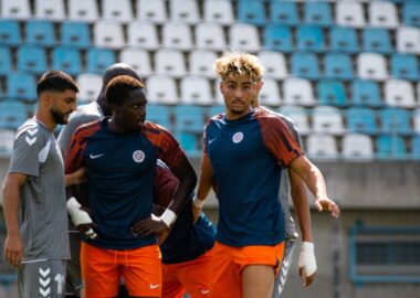 Ndiaye Meddah | AllezPaillade.com Three soccer players in blue and orange training kits stand together on a sunny stadium field during practice, with empty blue seats behind them.
