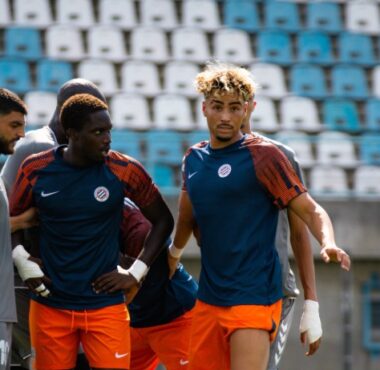 Three soccer players in blue and orange training kits stand together on a sunny stadium field during practice, with empty blue seats behind them.