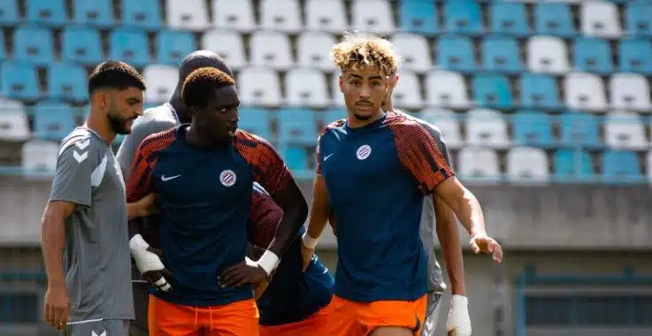 Three soccer players in blue and orange training kits stand together on a sunny stadium field during practice, with empty blue seats behind them.