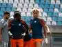 Three soccer players in blue and orange training kits stand together on a sunny stadium field during practice, with empty blue seats behind them.