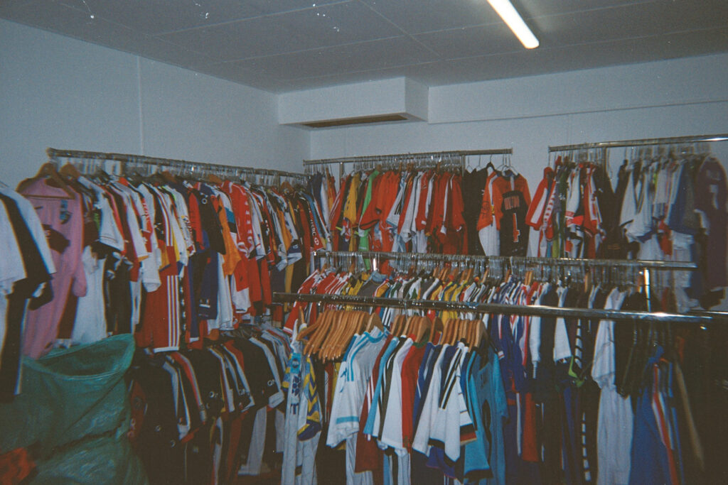 Photo06_3A | AllezPaillade.com Store interior with long racks of colorful sports jerseys hanging on metal rails under a bright ceiling light.