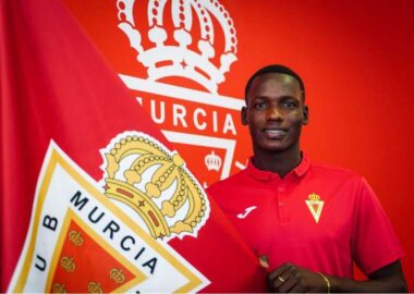 Sekou-Djanbou-2 | AllezPaillade.com Young man in a red polo shirt stands beside a large red flag with a crown crest, smiling.