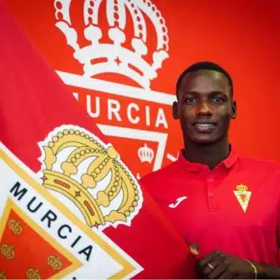 Young man in a red polo shirt stands beside a large red flag with a crown crest, smiling.