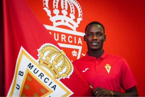 Young man in a red polo shirt stands beside a large red flag with a crown crest, smiling.