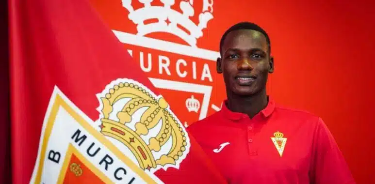 Sekou-Djanbou-2 | AllezPaillade.com Young man in a red polo shirt stands beside a large red flag with a crown crest, smiling.