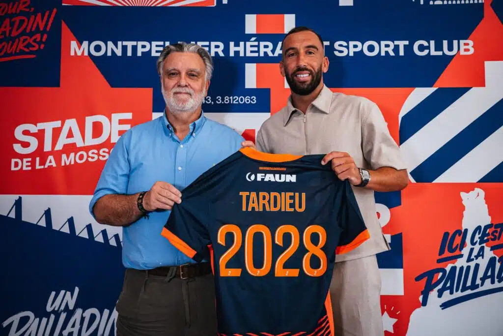 Tardieu | AllezPaillade.com Two men pose in front of a colorful backdrop, holding a navy and orange sports jersey with the number 2028 visible in orange.