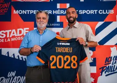 Two men pose in front of a colorful backdrop, holding a navy and orange sports jersey with the number 2028 visible in orange.
