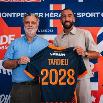 Two men pose in front of a colorful backdrop, holding a navy and orange sports jersey with the number 2028 visible in orange.
