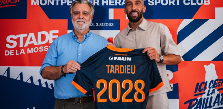 Tardieu | AllezPaillade.com Two men pose in front of a colorful backdrop, holding a navy and orange sports jersey with the number 2028 visible in orange.