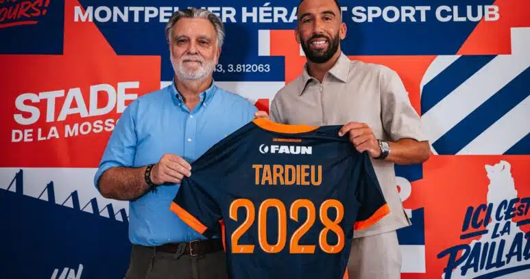 Two men pose in front of a colorful backdrop, holding a navy and orange sports jersey with the number 2028 visible in orange.