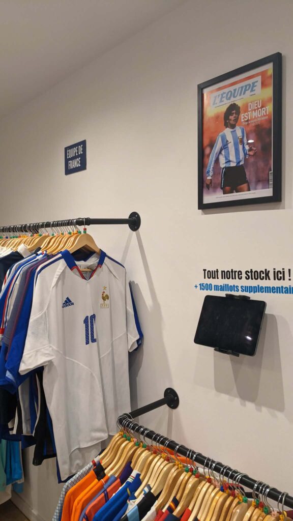 a82389bd-945d-465d-9300-e221ca975755 | AllezPaillade.com Store display with France national team jerseys hanging on a rack beside a framed poster on the wall, in a shop.