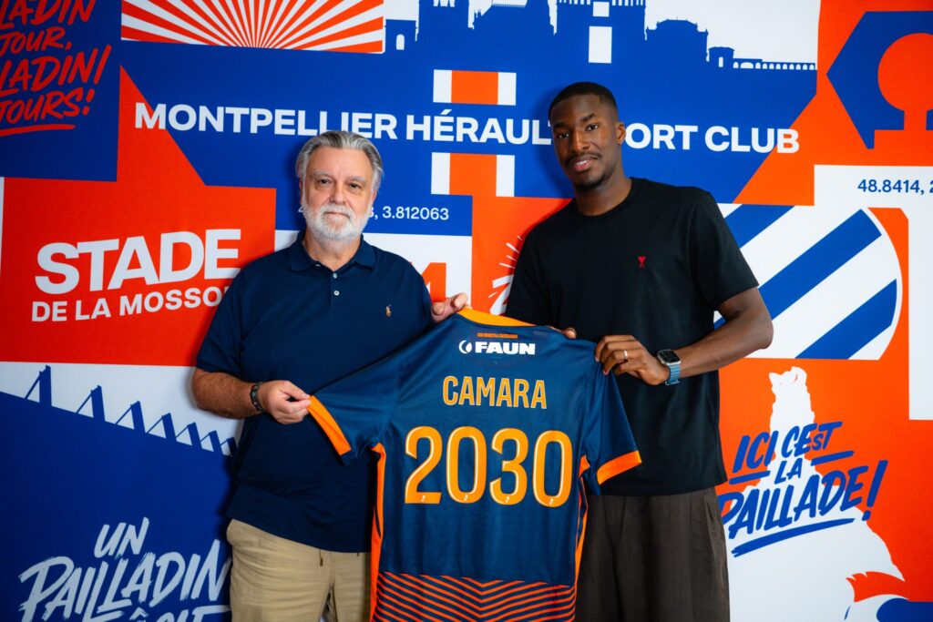 camara | AllezPaillade.com Two men pose together, one older with a blue polo and the other in a black shirt, holding a blue-and-orange jersey in front of a bold blue-orange mural.