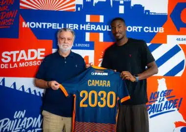 camara | AllezPaillade.com Two men pose together, one older with a blue polo and the other in a black shirt, holding a blue-and-orange jersey in front of a bold blue-orange mural.