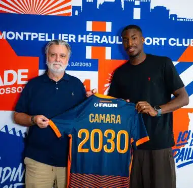 Two men pose together, one older with a blue polo and the other in a black shirt, holding a blue-and-orange jersey in front of a bold blue-orange mural.