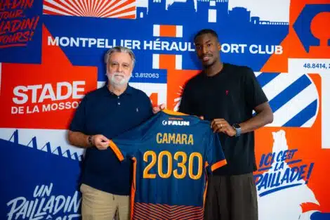Two men pose together, one older with a blue polo and the other in a black shirt, holding a blue-and-orange jersey in front of a bold blue-orange mural.
