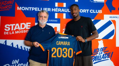 Two men pose together, one older with a blue polo and the other in a black shirt, holding a blue-and-orange jersey in front of a bold blue-orange mural.