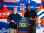 Two men pose together, one older with a blue polo and the other in a black shirt, holding a blue-and-orange jersey in front of a bold blue-orange mural.