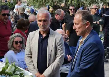 Two men in suits stand near a white flower arrangement at a memorial ceremony, surrounded by a crowd outdoors