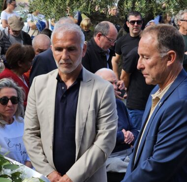 Two men in suits stand near a white flower arrangement at a memorial ceremony, surrounded by a crowd outdoors