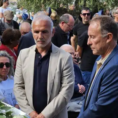 Two men in suits stand near a white flower arrangement at a memorial ceremony, surrounded by a crowd outdoors