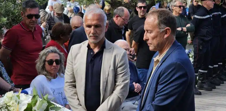 Two men in suits stand near a white flower arrangement at a memorial ceremony, surrounded by a crowd outdoors