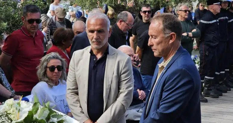 Two men in suits stand near a white flower arrangement at a memorial ceremony, surrounded by a crowd outdoors