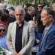Two men in suits stand near a white flower arrangement at a memorial ceremony, surrounded by a crowd outdoors
