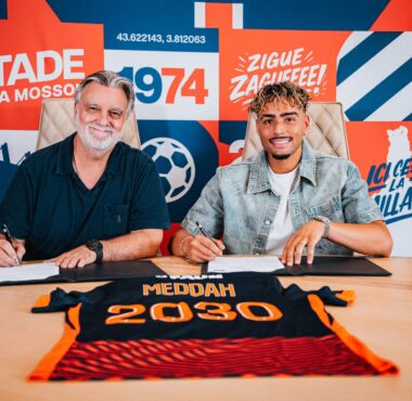 Two men sit at a table signing documents, with a bright graphic wall behind them.",